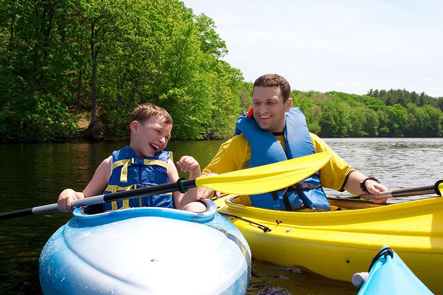 Employee Benefits - Happy Father and Son Wearing Safety Vests and Holding Paddles as They Laugh and Ride Their Canoes in the Lake