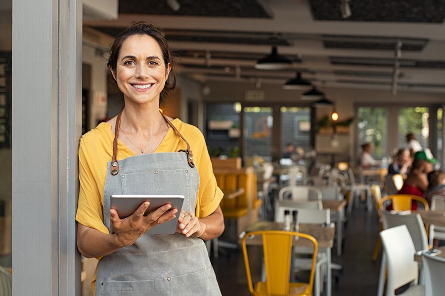 Specialized Business Insurance - Small Business Owner Wearing a Gray Smok and Yellow Shirt While Holding a Tablet at the Entrance of Her Shop